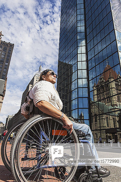 Frau im Rollstuhl schaut nach oben  während sie neben einem Wolkenkratzer mit reflektierender Glasfassade sitzt