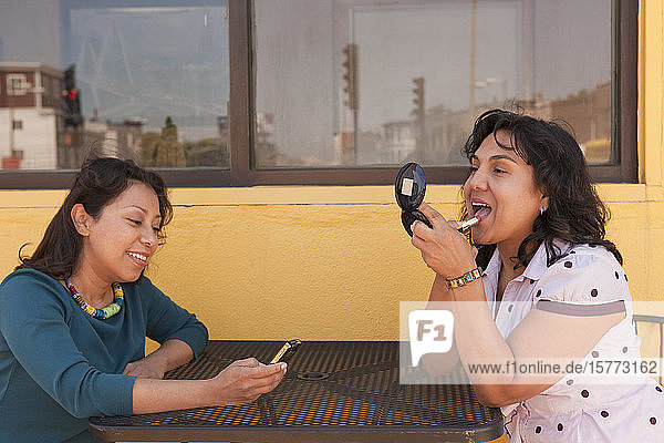Two young adult females sit at a table outside  one applying lipstick and the other on her smart phone