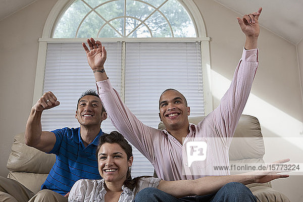 Drei junge Erwachsene sitzen in einem Wohnzimmer zu Hause und sehen jubelnd fern