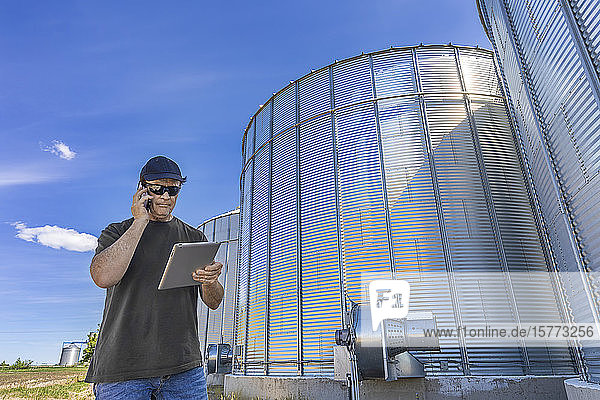 A farmer stands beside grain storage bins using a cell phone and holding a tablet; Alberta  Canada