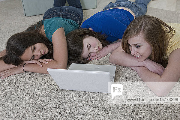 Drei Mädchen im Teenageralter benutzen einen Laptop auf dem Teppichboden  zwei schlafen und eine schaut auf den Bildschirm