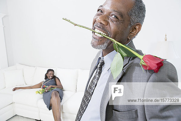Porträt eines Mannes mittleren Alters  der eine Rosenblüte zwischen seinen Zähnen hält  mit einer lächelnden Frau mittleren Alters im Hintergrund