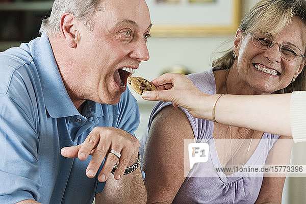 Mature man eating a cookie with a mature woman in the background.