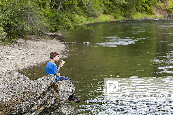 A teenage boy sits on a rock ledge over a river eating a sandwich for lunch; British Columbia  Canada