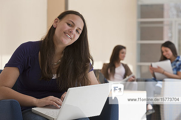 Teenager-Mädchen  das in der Schule einen Laptop benutzt  mit zwei Schülern  die im Hintergrund auf Lehrbücher schauen