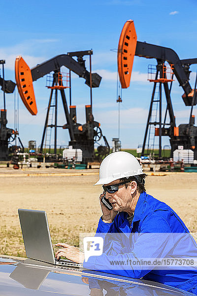 Man working on a laptop and smart phone with pumpjacks in the background; Alberta  Canada