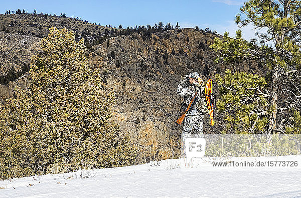 Hunter with camouflage clothing and rifle looking out with binoculars; Denver Colorado United States of America