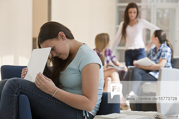 Teenage girl using laptop at school and hiding the screen from others with two students looking at textbooks in the background