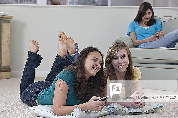 Drei Mädchen im Teenageralter sitzen zusammen zu Hause  zwei sehen fern und wechseln den Kanal mit der Fernbedienung  die andere sitzt in einem Sessel und hört Musik
