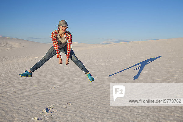 A young woman leaps in the air on the white sand with blue sky White Sands National Monument; Alamogordo New Mexico United States of America