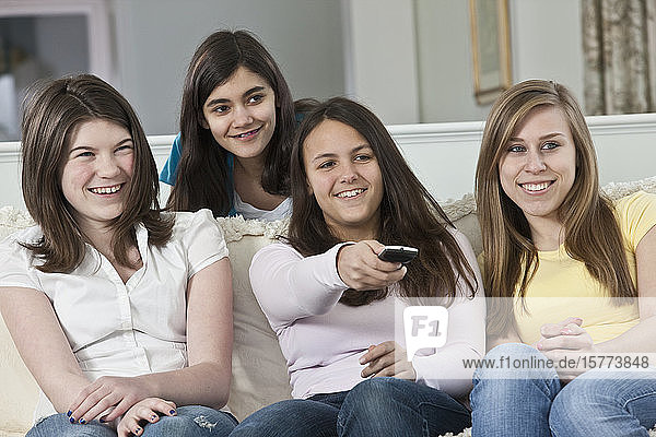 Four teenage girls sitting on a couch at home watching TV and changing the channel with the remote control