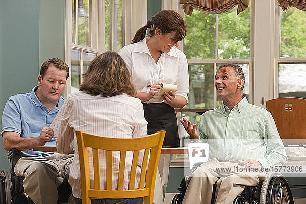 Eine Kellnerin nimmt die Bestellung einer Mahlzeit von einer Gruppe an einem Tisch auf  darunter zwei Männer im Rollstuhl