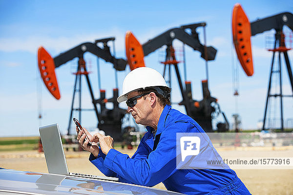 Man working on a laptop and smart phone with pumpjacks in the background; Alberta  Canada