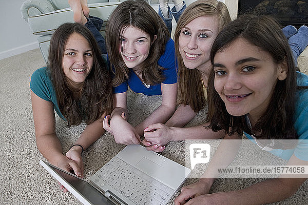 Vier Mädchen im Teenageralter benutzen einen Laptop  während sie auf dem Teppichboden liegen und in die Kamera schauen