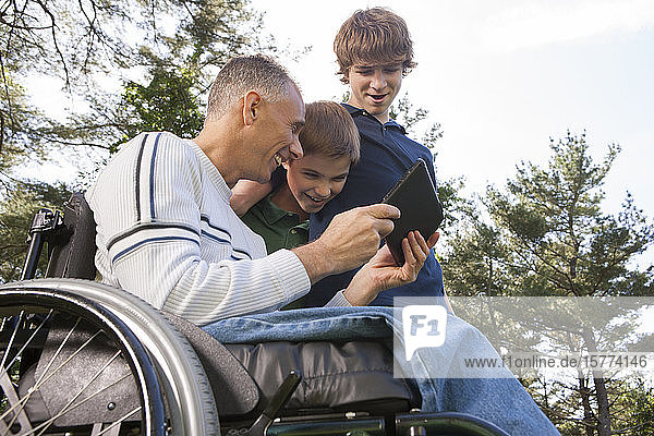 Ein Mann im Rollstuhl teilt einen Moment mit seinen Söhnen  während sie etwas auf einem Tablet anschauen und zusammen lachen