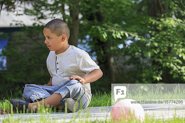 Pre-teen boy outside sitting on the ground with trees in the background
