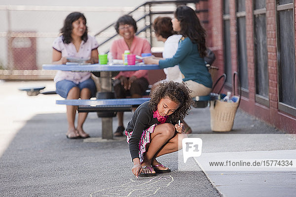 Ein Mädchen hockt auf dem Bürgersteig und zeichnet mit Kreide ein Himmelfahrtskommando  während drei Frauen hinter ihr sitzen und sich unterhalten