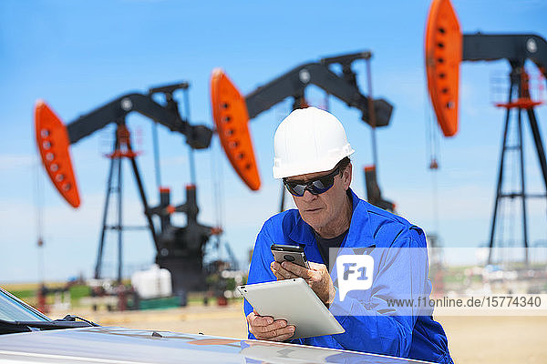 Man working on a tablet and smart phone with pumpjacks in the background; Alberta  Canada