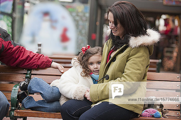 Eine kleine Tochter ruht sich auf ihrer Mutter auf einer Bank in einer Einkaufszone aus