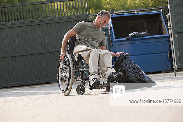 Mann im Rollstuhl hebt einen Müllsack mit einer Mülltonne im Hintergrund