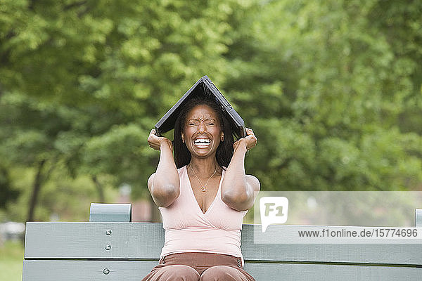 Mid adult woman sitting on a bench and holding a laptop over her head