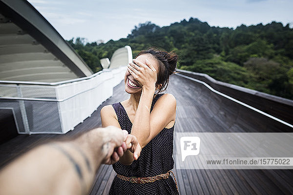 Lächelnde junge Frau steht auf einer Brücke  bedeckt ihr Gesicht und hält die Hand eines Mannes.