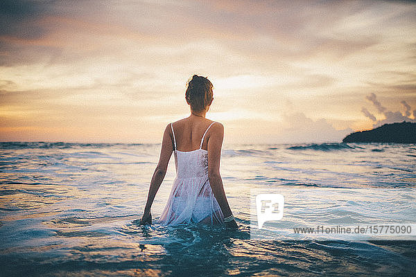 Rückansicht einer jungen Frau in einem weißen Kleid  die bei Sonnenuntergang im Meer spazieren geht.