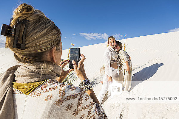 Eine Frau fotografiert ihre Kinder mit einem Smartphone in einer weißen Sanddünenlandschaft unter blauem Himmel.