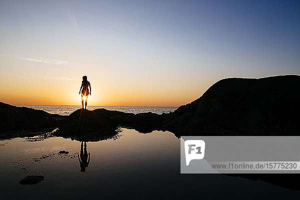 Silhouette of woman standing on a rock by the ocean at sunset.