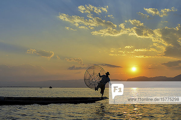 Traditioneller Fischer  der auf einem Bein auf einem Boot balanciert  einen Angelkorb hält  beim Angeln auf dem Inle-See bei Sonnenuntergang  Myanmar.
