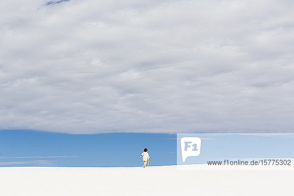 Ein kleiner Junge  der in weißen Sanddünen  einem Streifen blauen Himmels am Horizont und dichten Wolken spazieren geht.