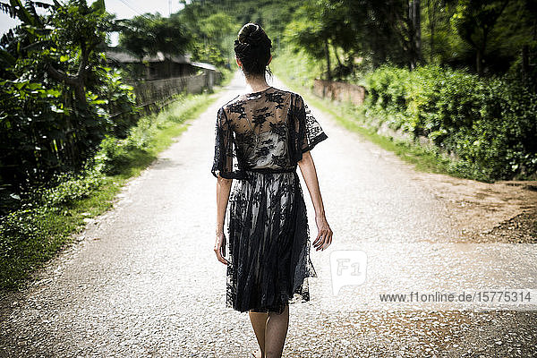 Rückansicht einer Frau in einem schwarzen Spitzenkleid  die eine ländliche Landstraße entlanggeht.