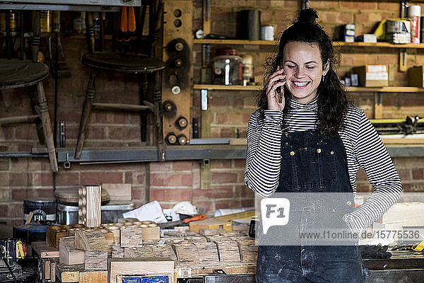 Frau mit langen braunen Haaren in Latzhose in Holzwerkstatt stehend  spricht mit dem Handy.