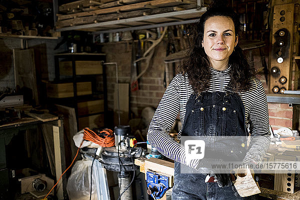 Woman with long brown hair wearing dungarees standing in wood workshop  smiling at camera.