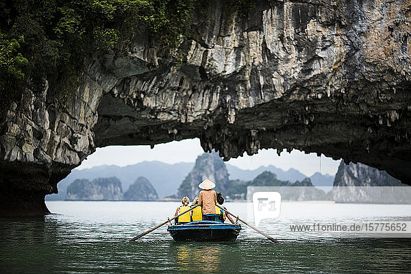 Rear view of man wearing straw hat transporting small group of people on a boat  rowing underneath natural rock arch.