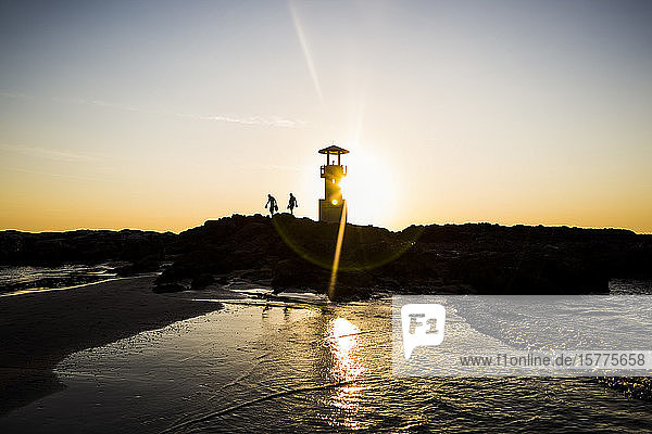 Silhouette of two people walking past lighthouse by the ocean at sunset.