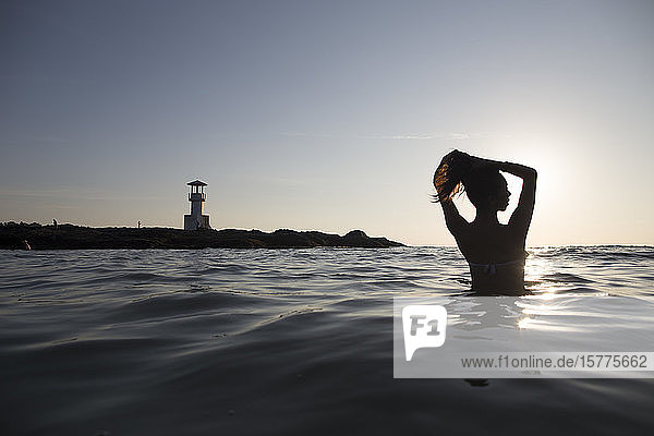 Rückansicht einer im Meer badenden Frau bei Sonnenuntergang  in der Ferne Leuchtturm.