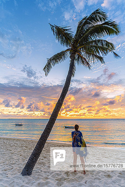 Rückansicht einer Frau mit Sarong  die den Ozean bei Sonnenuntergang am palmengesäumten Strand bewundert  Le Morne Brabant  Black River  Mauritius  Indischer Ozean  Afrika