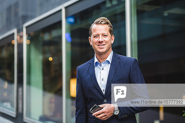 Portrait of smiling mature businessman with smart phone standing against office building