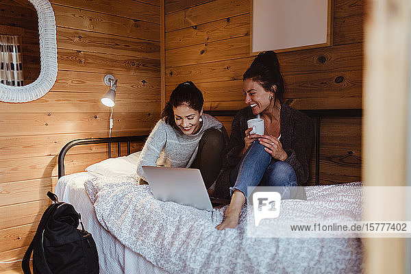 Smiling female friends using laptop while sitting on bed at home