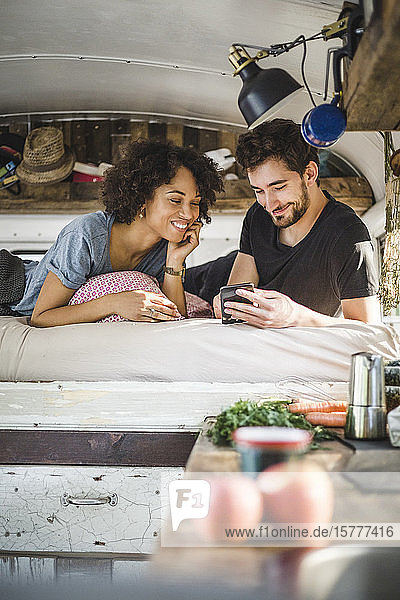 Lächelndes Paar  das sich ein Smartphone teilt  während es im Wohnwagen auf dem Bett liegt