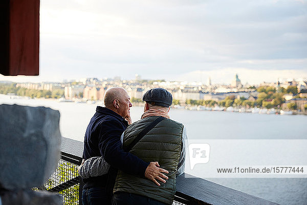 Rear view of gay couple with arm around standing against railing by river in city