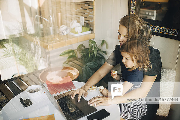 Mutter benutzt Laptop  während die Tochter auf dem Schoß sitzt und durch ein Glasfenster gesehen wird