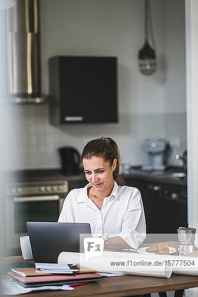 Lächelnde Freiberuflerin benutzt Laptop  während sie zu Hause am Tisch sitzt