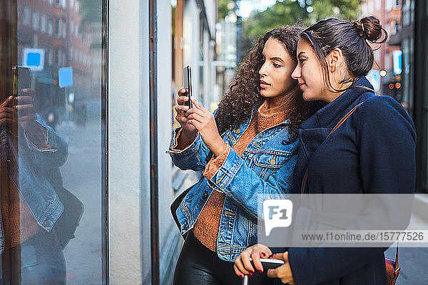 Female friends photographing store window with smart phone in city