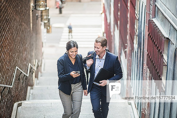 Smiling male and female entrepreneurs using phone while climbing staircase in city