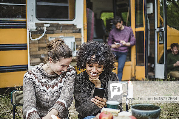 Lächelnde Freundinnen teilen sich ein Smartphone  während sie beim Camping gegen den Wohnwagen sitzen