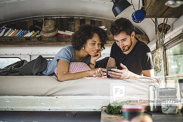 Junges Paar  das sich ein Smartphone teilt  während es im Wohnwagen auf dem Bett liegt