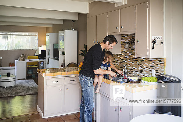 Vater wäscht Küchenutensilien,  während die Tochter auf dem Küchentisch sitzt