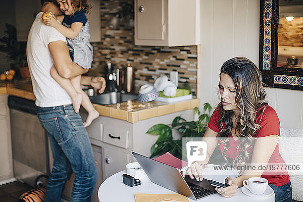 Mother using laptop while father carrying daughter and working in domestic kitchen